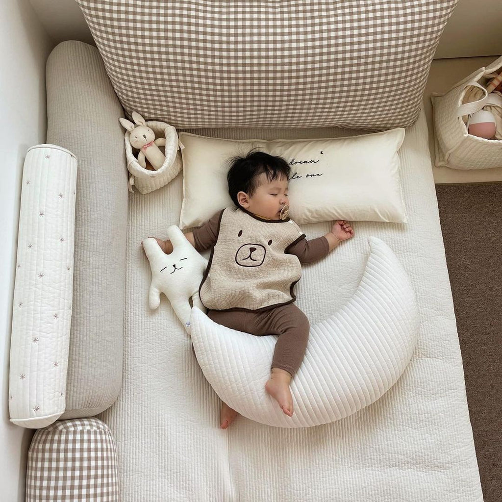 Bébé dormant paisiblement avec le coussin lune Be'Lune blanc, dans un lit douillet décoré d’accessoires en coton naturel.
